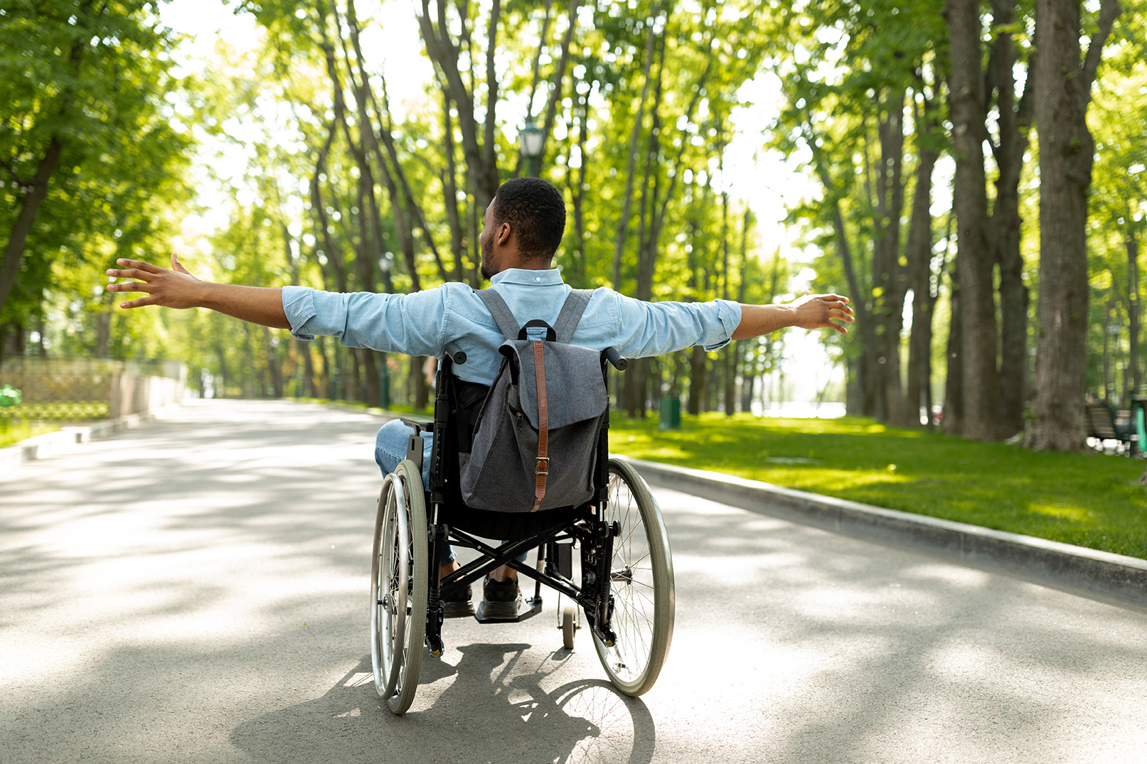 Back view of young disabled black man in wheelchair spreading his arms, feeling happy and free on walk at city park. Full length of handicapped guy with backpack going on trip outdoors