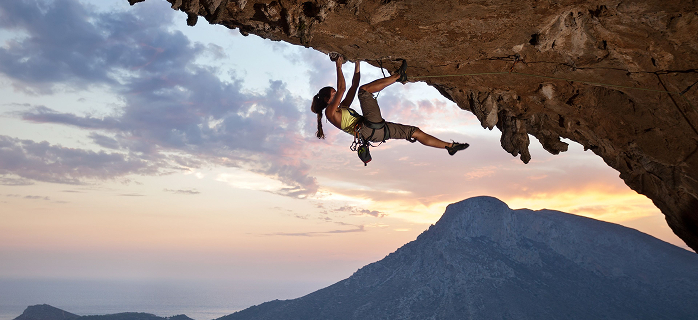 Female rock climber gripping an overhanging cliff face at sunset with a mountain landscape in the background, representing physical strength and recovery
