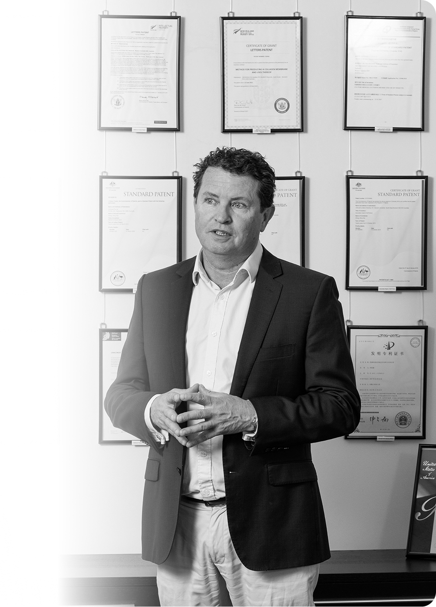 Orthocell executive in a dark blazer standing in front of a wall displaying multiple framed international patents and certificates of grant