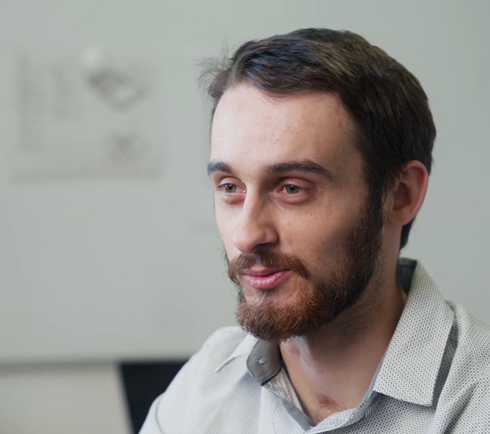 Young man with a beard speaking during an interview in a modern office setting with a whiteboard in the background