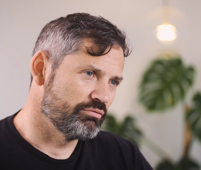 Middle-aged man with a grey beard speaking during an interview in a bright room with an indoor plant in the background