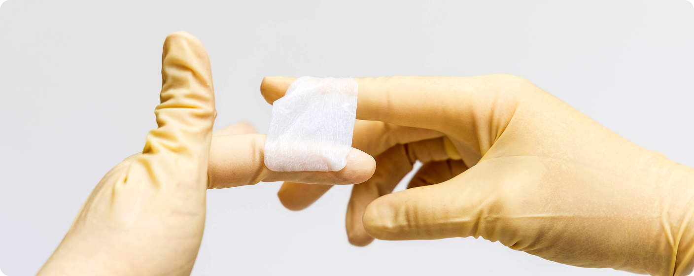 Gloved hands handling a small translucent collagen medical device membrane in a sterile clinical environment
